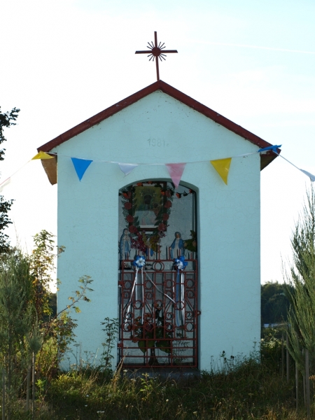 Biała kapliczka z czerwoną dachówką i metalowym krzyżem na szczycie, ozdobiona kolorowymi chorągiewkami, otoczona zielenią i niską roślinnością.