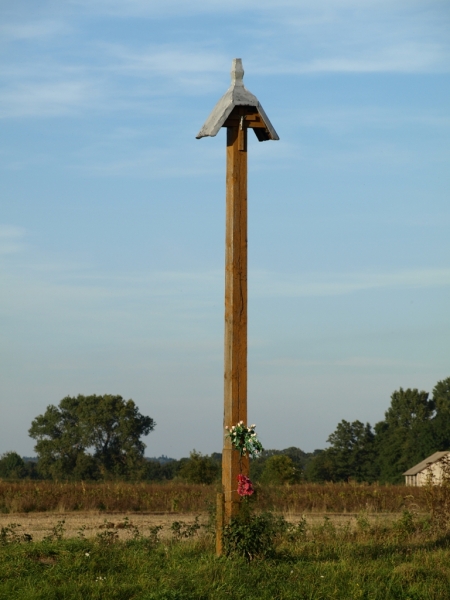 Wysoki drewniany krzyż z daszkiem, przywiązane kwiaty, zielona trawa, w tle drzewa i jasne niebo.