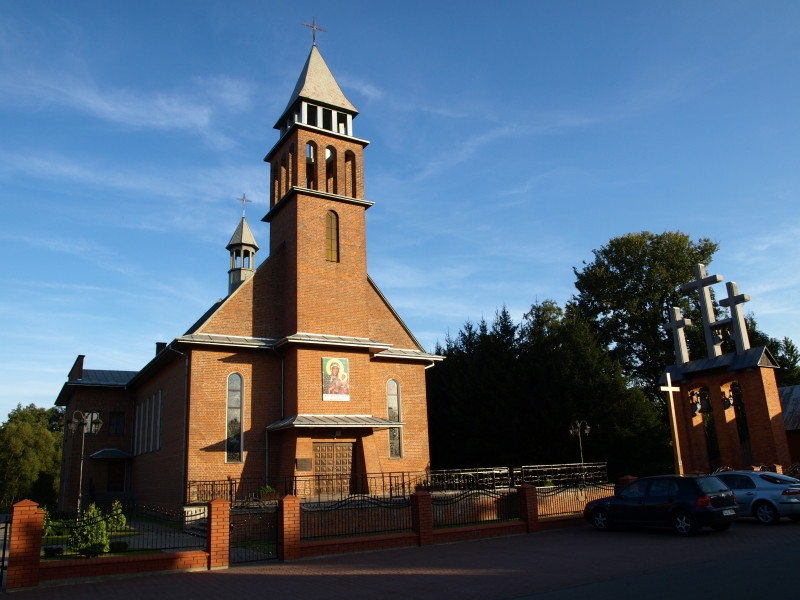 Ceglana czerwona wieża kościoła z dwoma krzyżami na dachu, obrazem świętej postaci, ogrodzeniem i trzema krzyżami na osobnym, ceglastym filarze, samochody i drzewa w tle.