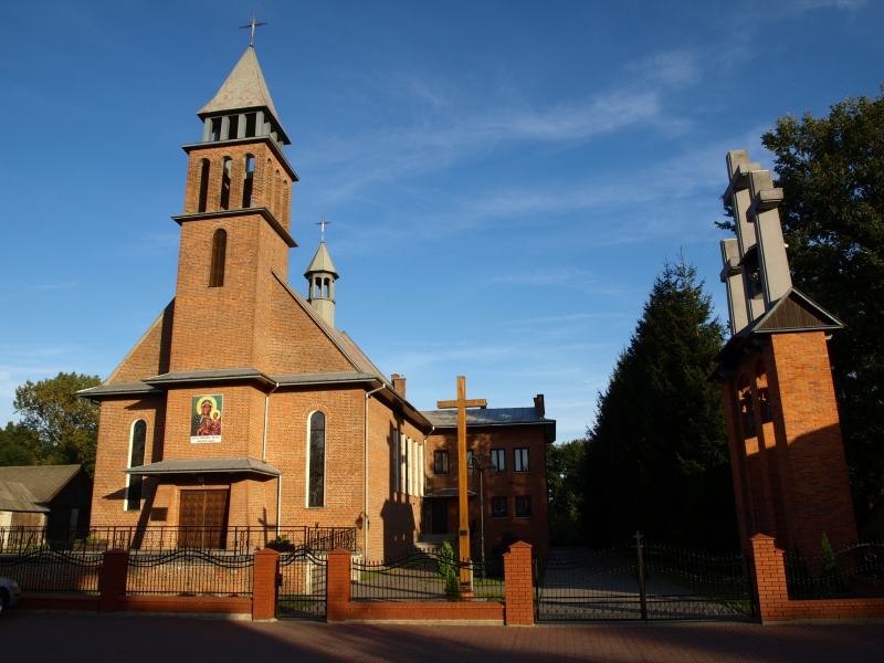 Ceglany kościół z wysoką wieżą, krzyżem przed budynkiem i dzwonnicą po prawej stronie, na tle błękitnego nieba i drzew.