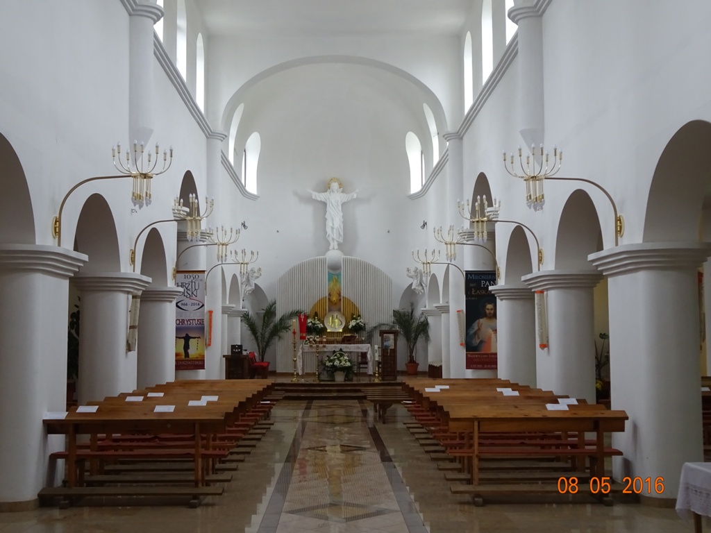 Wnętrze jasnego kościoła z białymi ścianami, łukowatymi kolumnami, drewnianymi ławkami i dużą białą figurą Chrystusa nad ołtarzem.