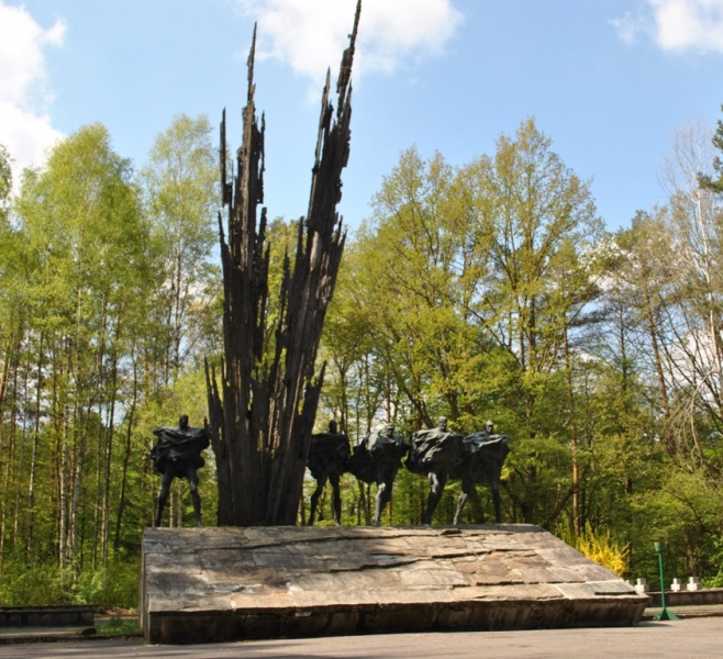Pomnik z czterema postaciami na cokole i wysokim, spiczastym, czarnym elementem w centrum, otoczony zielonymi drzewami pod niebieskim niebem.