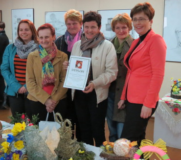Grupa sześciu kobiet w kolorowych ubraniach stoi za stołem z dekoracjami i kwiatami, jedna kobieta trzyma dyplom. W tle rysunki na ścianie.