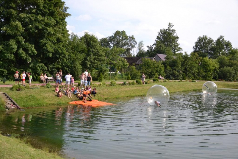 Grupa ludzi nad brzegiem stawu z zieloną trawą i drzewami, dwie przezroczyste kule wodne z osobami w środku na wodzie.