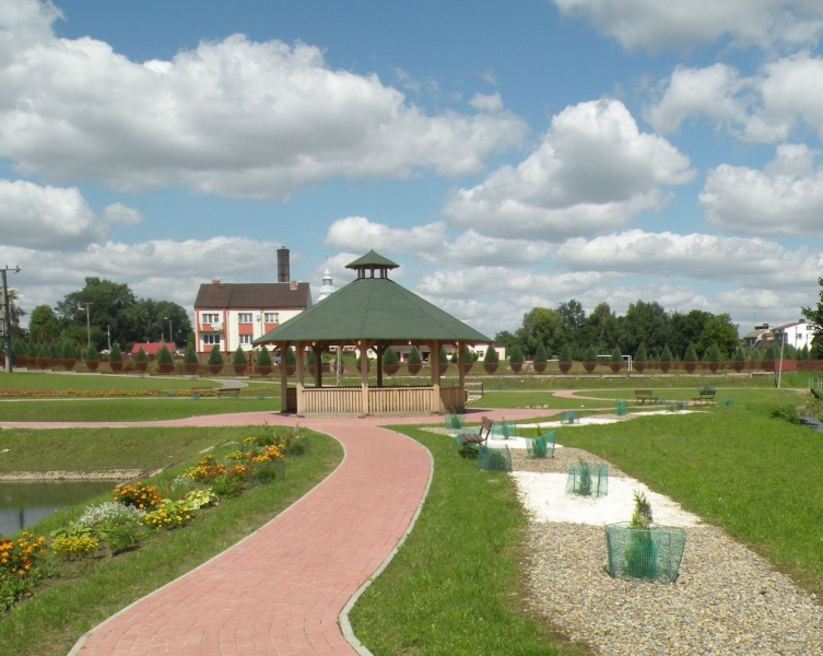 Park z czerwoną ścieżką, drewniana altana z zielonym dachem, ławki, kwiaty i trawa, w tle domy i niebieskie niebo z białymi chmurami.