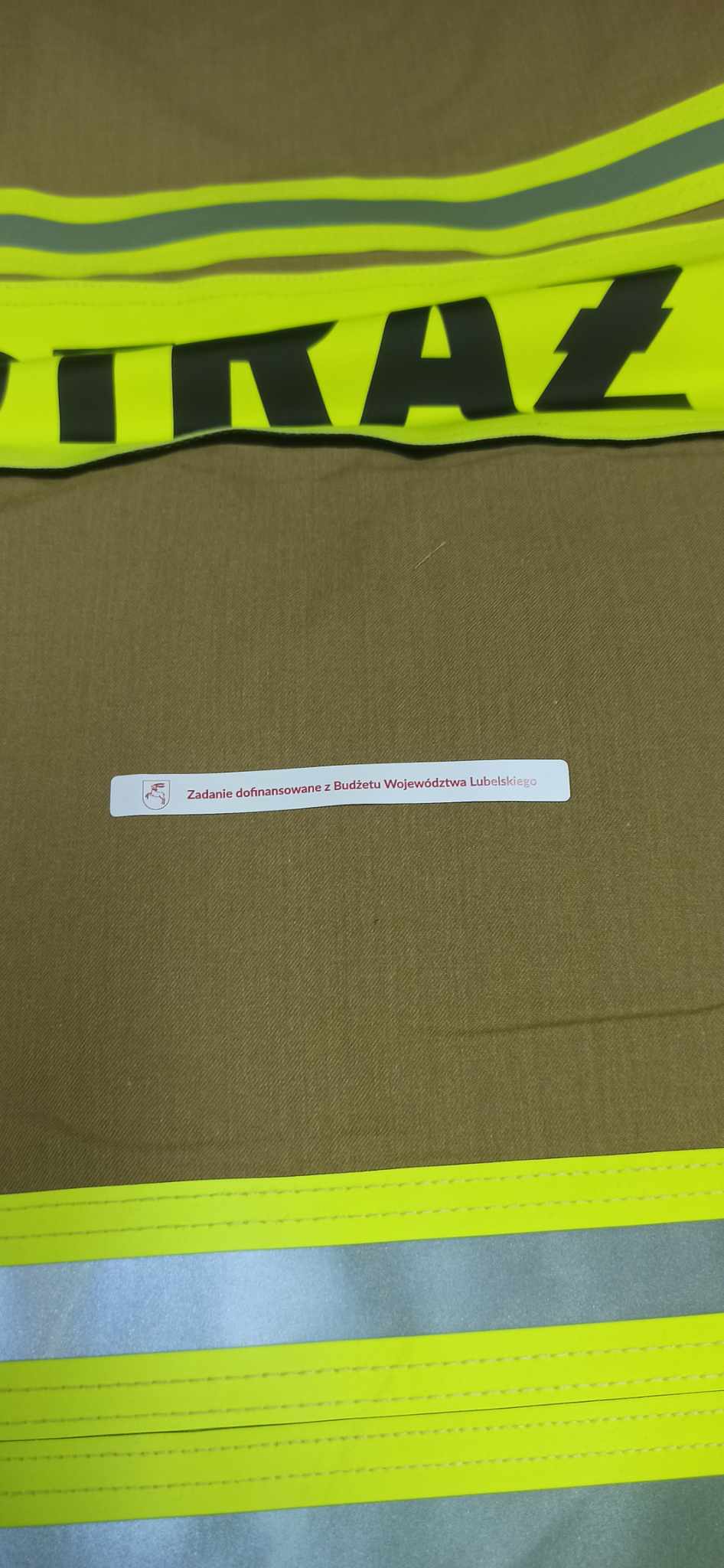 Brązowy materiał z żółtymi odblaskowymi pasami, na którym znajduje się biały napis z czerwonym tekstem i małym herbem.