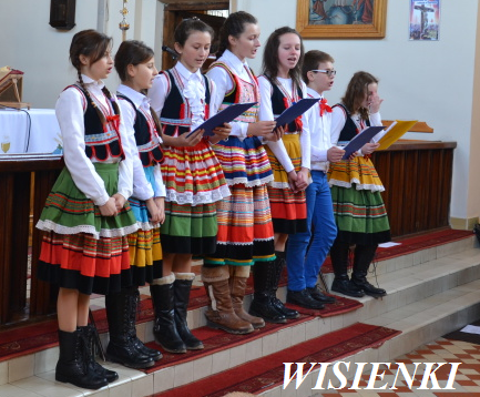 Sześć dzieci w tradycyjnych, kolorowych strojach ludowych stoi na schodach w pomieszczeniu z drewnianą balustradą i białymi ścianami, trzymając teczki z papierami.