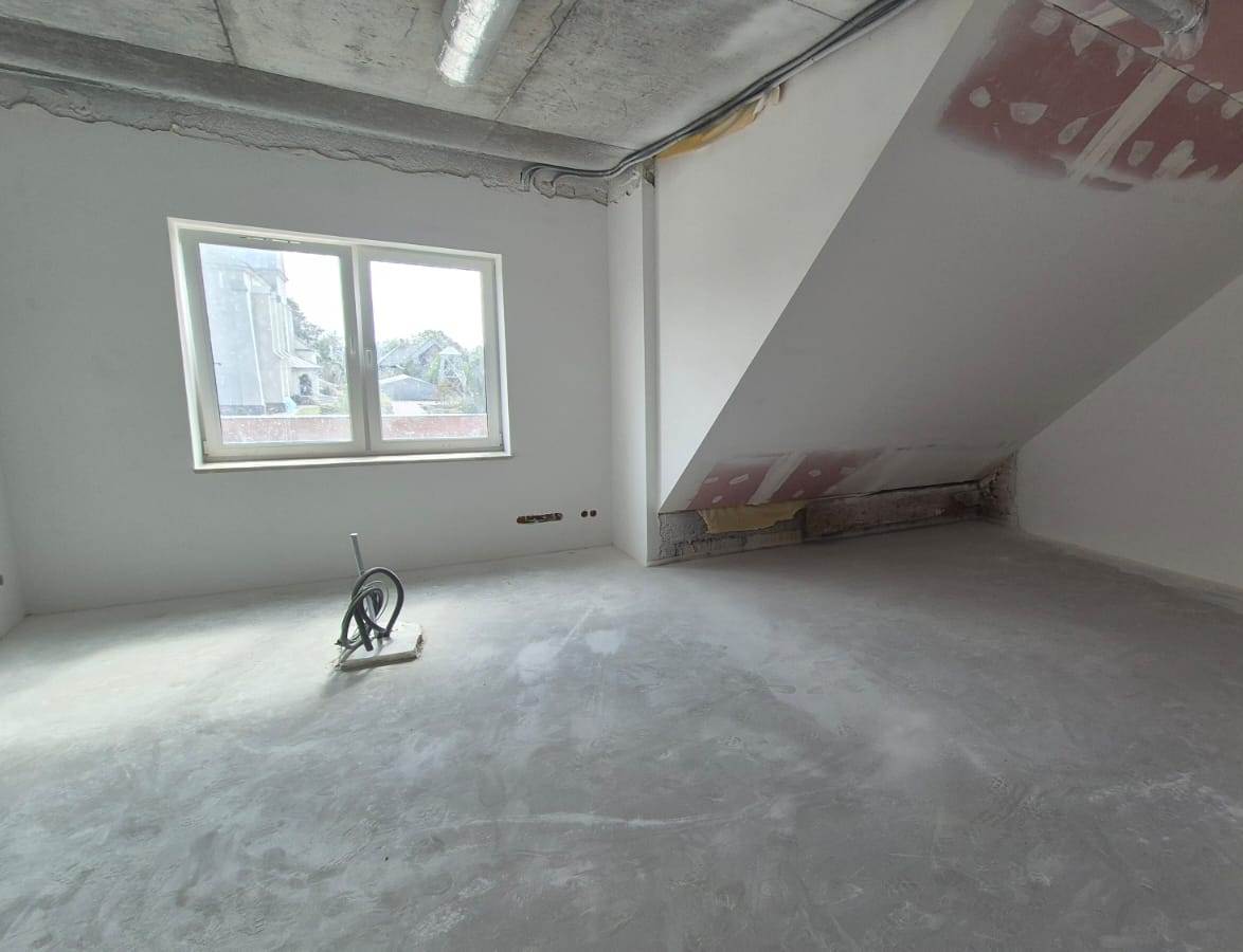 Pusta, jasna, nieukończona poddaszowa przestrzeń z betonową podłogą, białymi ścianami, skośnym sufitem, oknem oraz widocznymi rurami i przewodami.