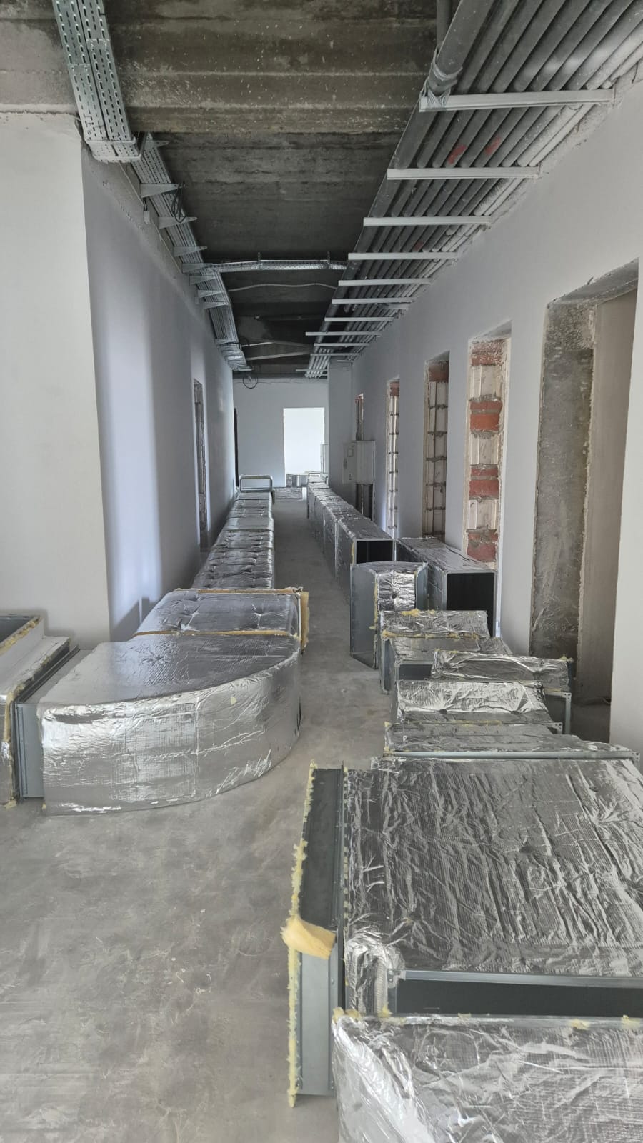 Korytarz z surową podłogą, białe ściany, sufit betonowy z metalowymi korytkami kablowymi, na podłodze metalowe, częściowo izolowane kanały wentylacyjne.
