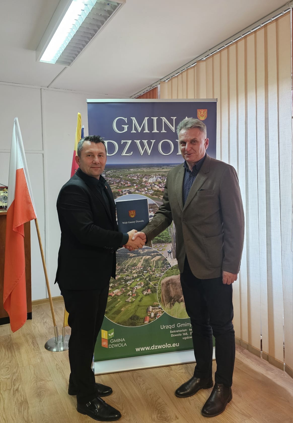 Dwóch mężczyzn w garniturach podaje sobie ręce na tle banera z napisem "Gmina Dzwola" i flag Polski oraz innej na stojakach.
