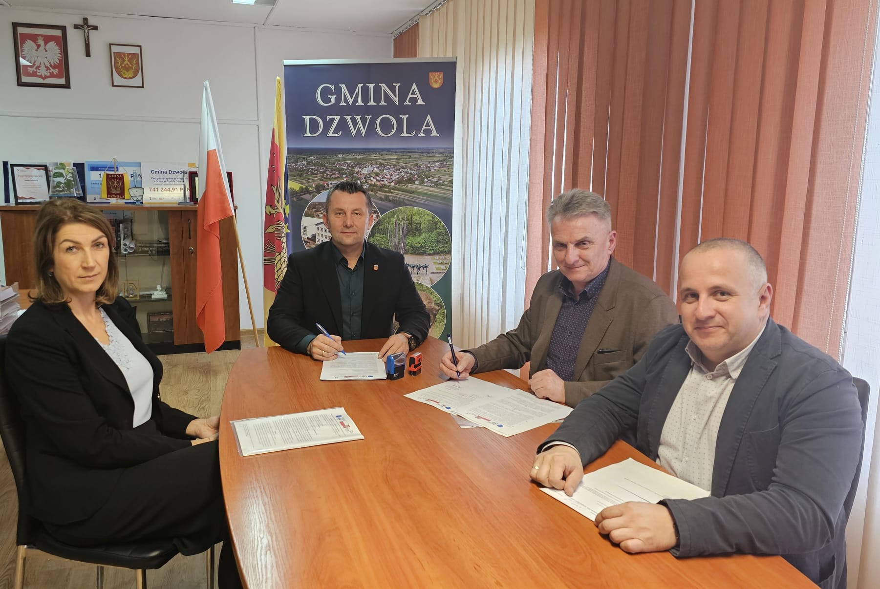 Cztery osoby siedzące przy drewnianym stole, na stole dokumenty, w tle flagi Polski i gminy, baner z napisem "Gmina Dzwola", jasne zasłony, jasne ściany.
