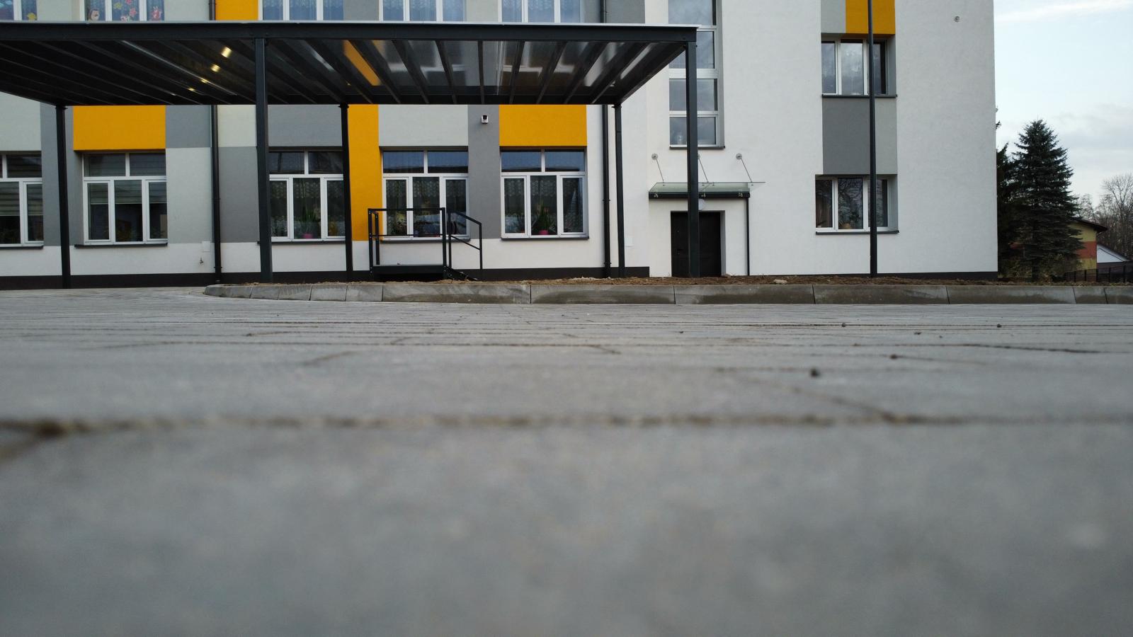 Szare płyty chodnikowe na pierwszym planie, za nimi nowoczesny budynek z białą, szarą i żółtą elewacją, szklany daszek nad wejściem, metalowa konstrukcja zadaszenia.