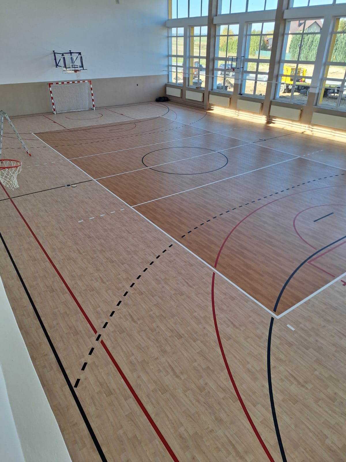 Drewniana podłoga hali sportowej z pomalowanymi liniami do różnych dyscyplin, okna na całej ścianie, bramka do piłki ręcznej i kosz do koszykówki.
