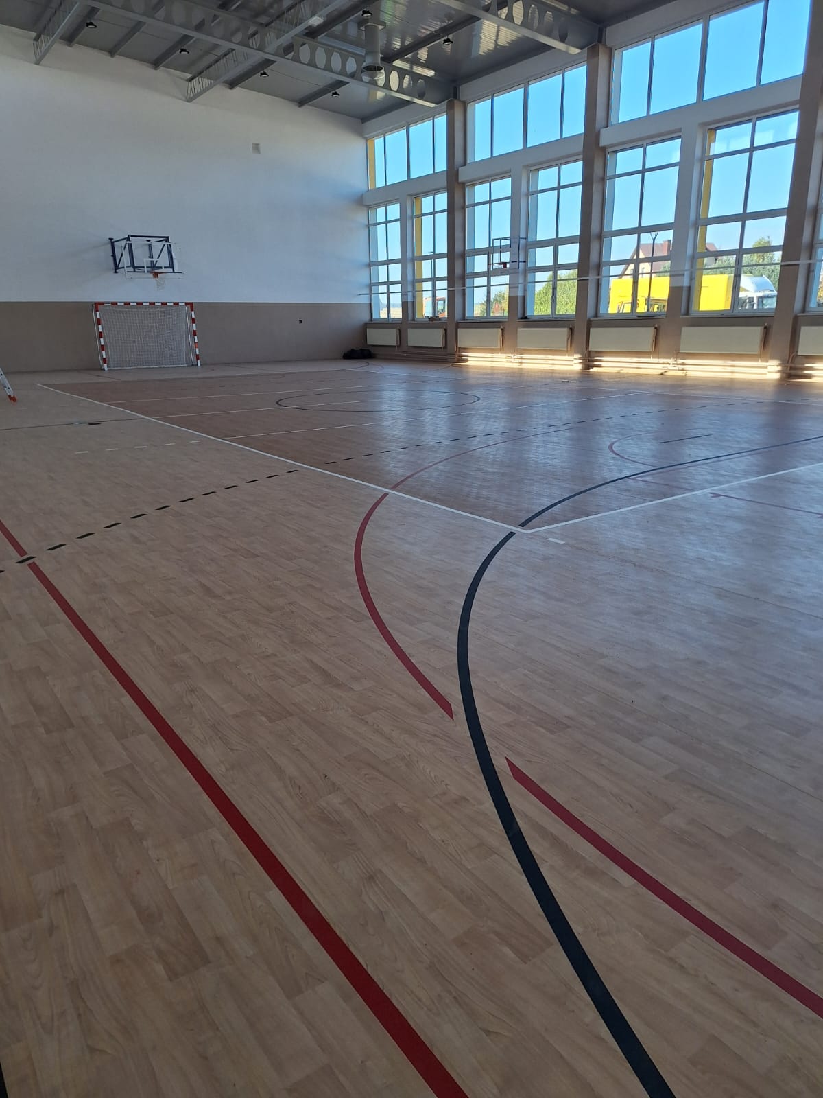 Duża hala sportowa z jasną drewnianą podłogą, pomalowanymi liniami do różnych gier, wysokimi oknami i koszem do koszykówki nad bramką do piłki ręcznej.