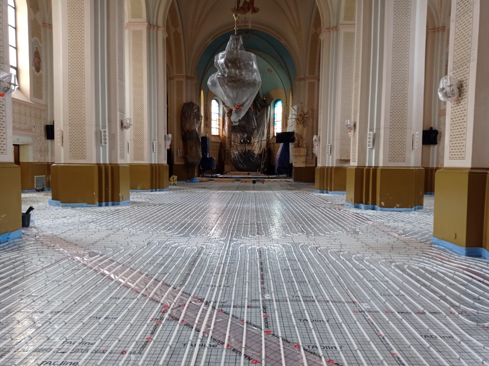 Wnętrze dużej przestrzeni z wysokim sklepieniem, kolumny białe z brązową dolną częścią, na podłodze białe rury ułożone w równoległe i zakrzywione linie, elementy przykryte folią.