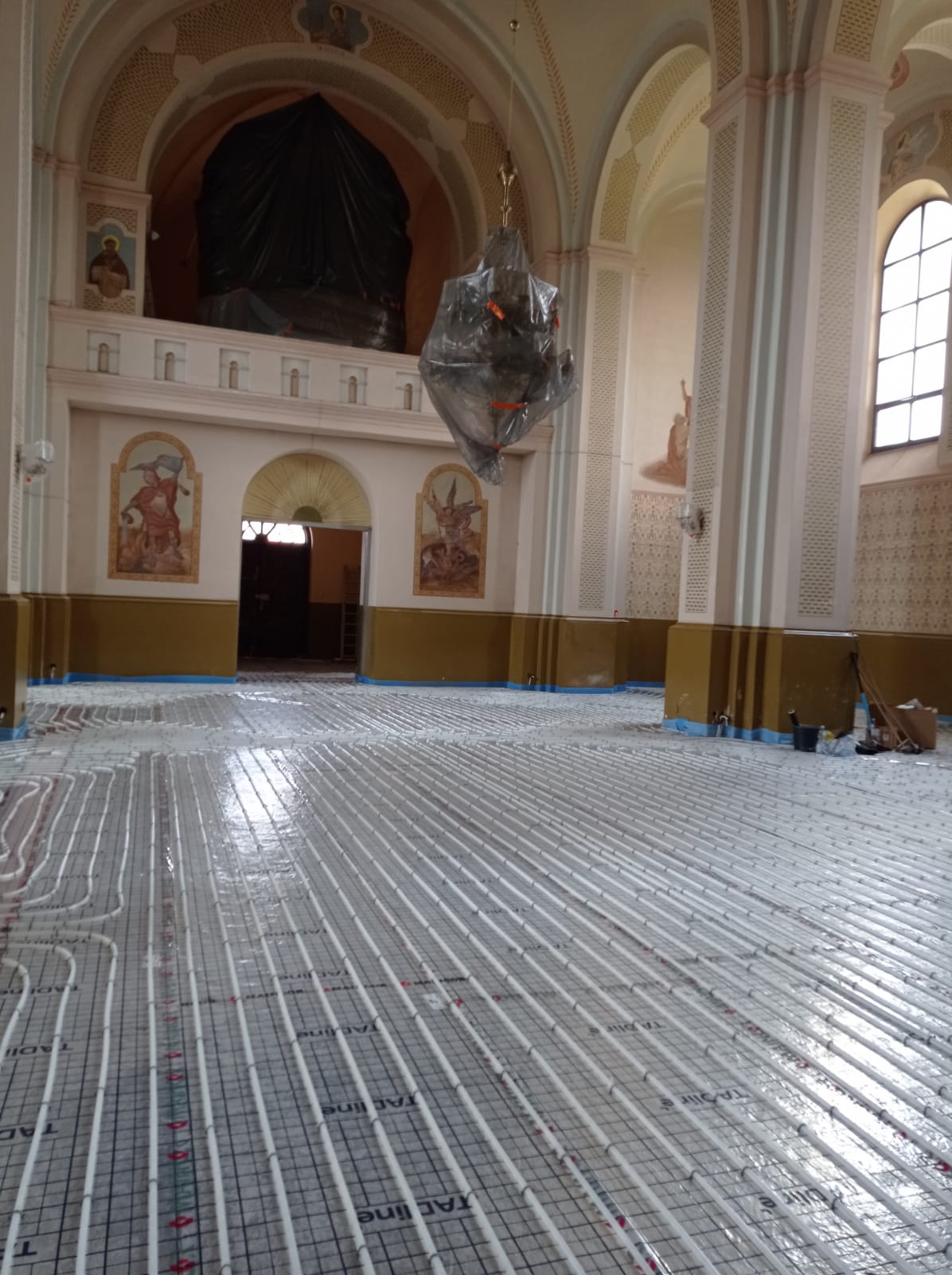 Wnętrze zabytkowego budynku z wysokimi łukowymi sufitami, z instalacją rur na podłodze, ściany w dolnej części pomalowane na oliwkowo, górna część jasna z malowidłami.