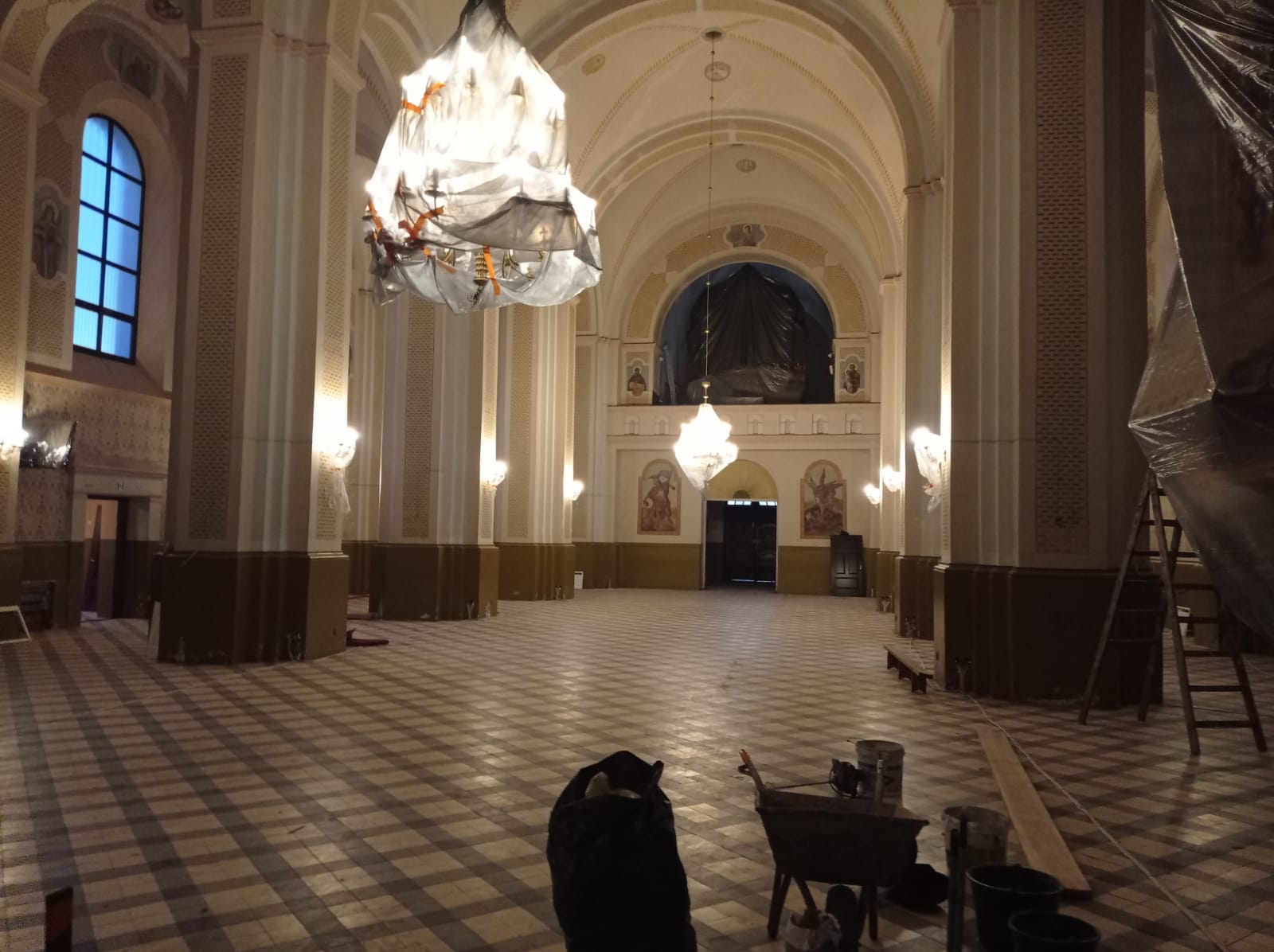 Wnętrze dużej hali z wysokim sklepieniem, jasnobeżowe ściany z ornamentami, okna z niebieskim światłem, podłoga w kratę, wiszące żyrandole przykryte folią, narzędzia i sprzęty budowlane na podłodze.