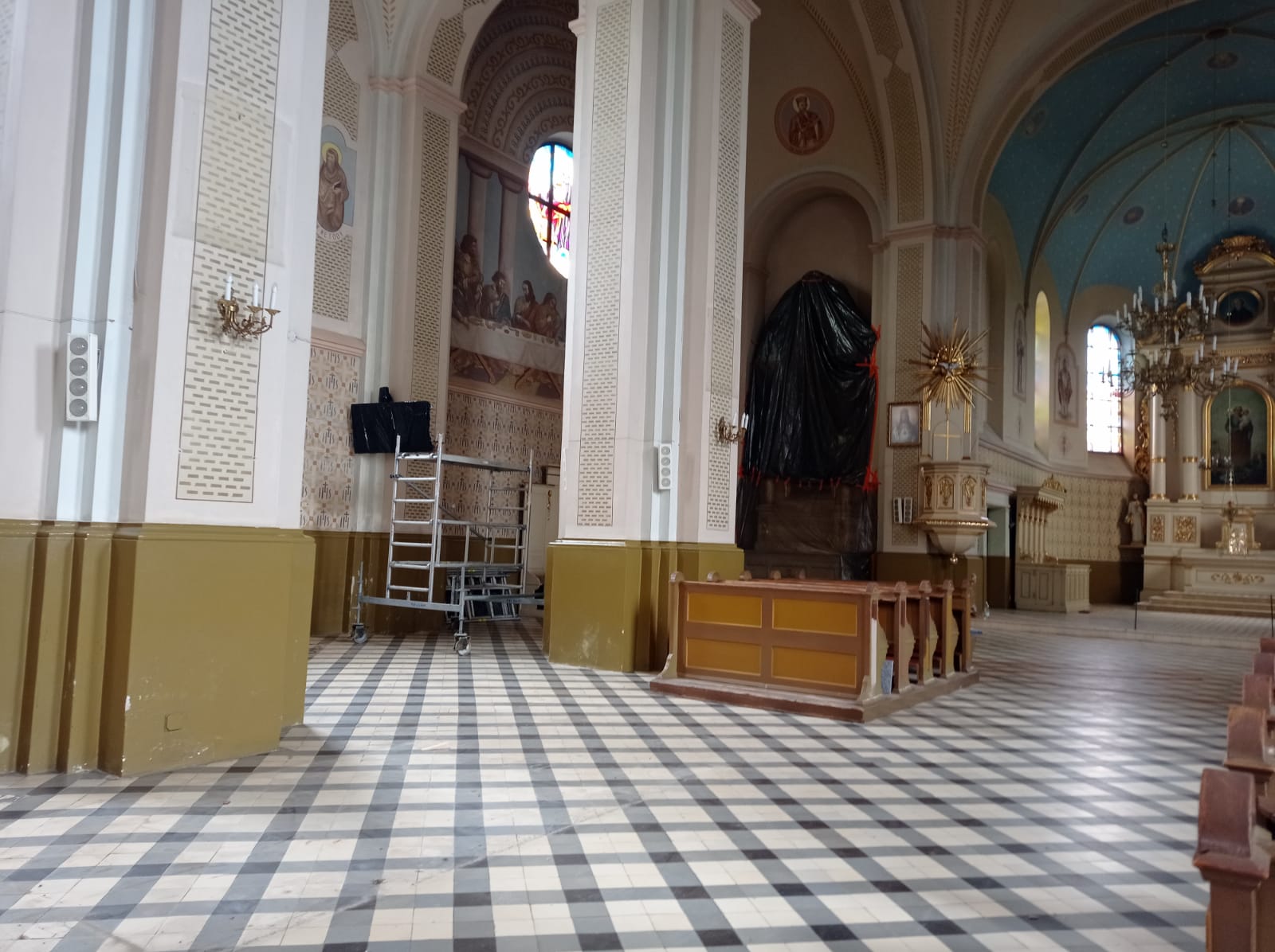 Wnętrze kościoła z białymi i oliwkowymi filarami, czarno-białą szachownicą na podłodze, ławkami, złoconym ołtarzem, rusztowaniem i czarną folią na jednej z nisz.