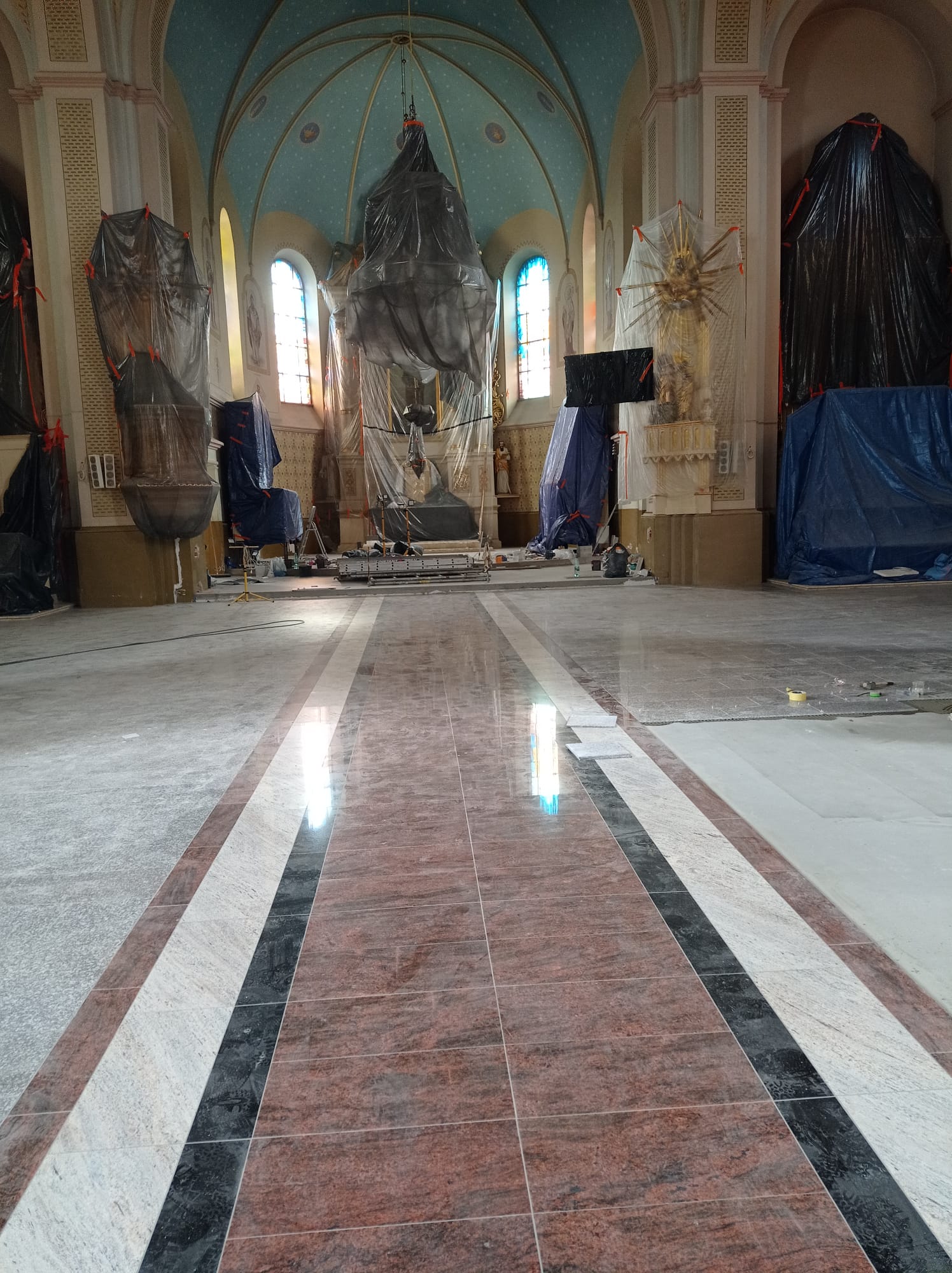 Wnętrze kościoła z marmurową podłogą i łukowym sklepieniem, ołtarz i figury przykryte czarnymi i przezroczystymi foliamii, światło wpada przez kolorowe witraże.