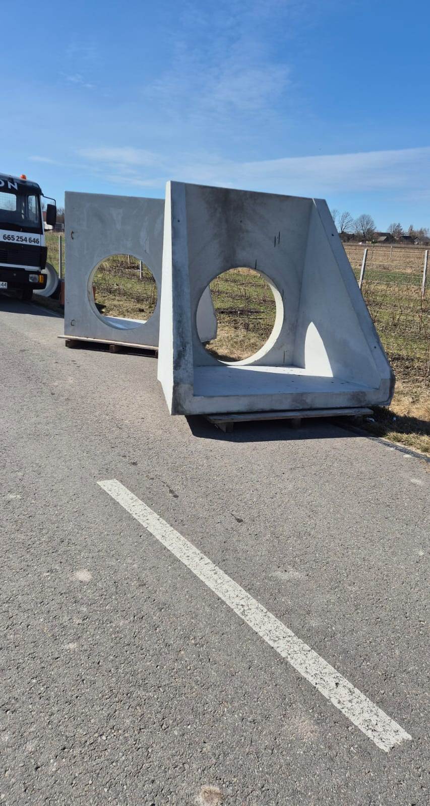 Dwa duże, szare betonowe prefabrykaty z okrągłymi otworami, ustawione na paletach na asfalcie przy polu, w tle częściowo widoczna ciężarówka.