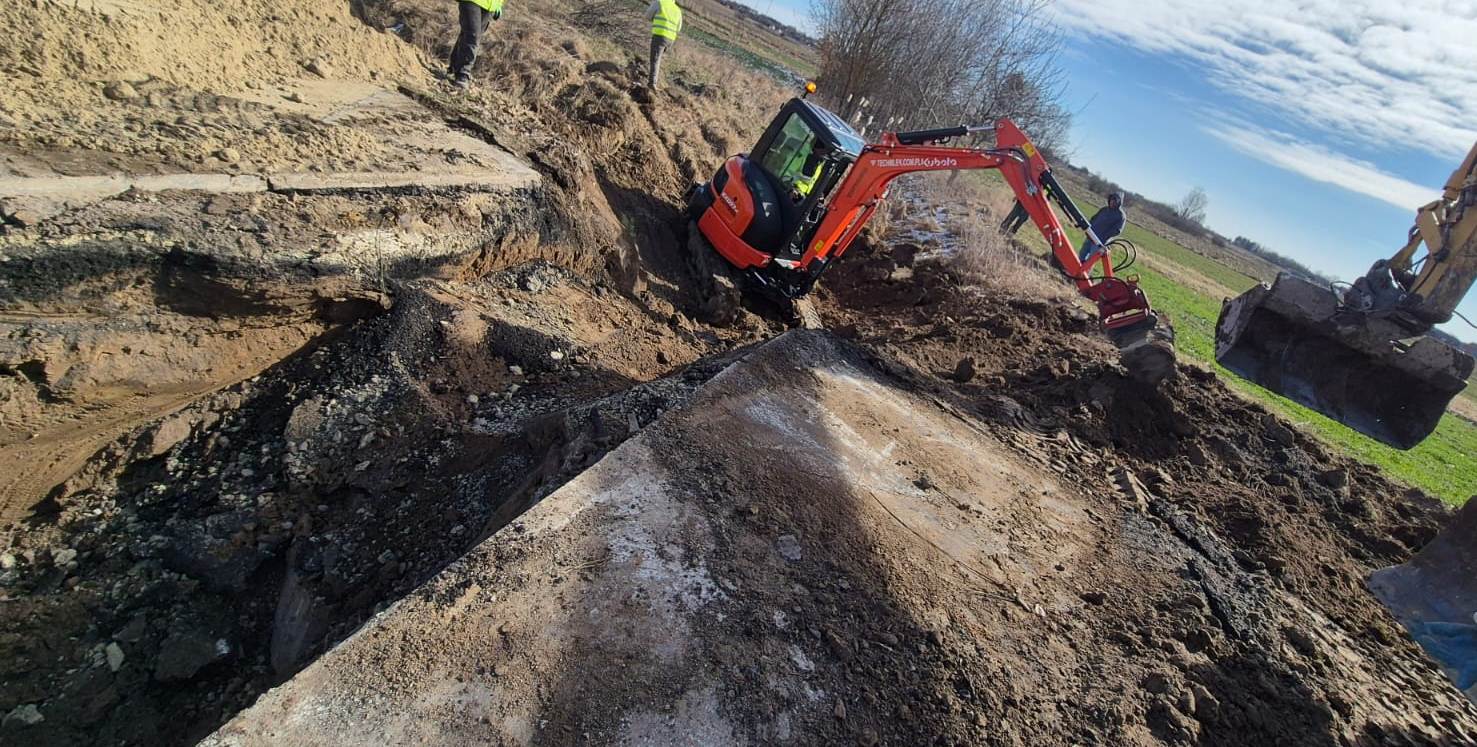 Koparka w kolorze pomarańczowym i żółtym kopiąca ziemię na polu, widoczne fragmenty betonu, kilka osób w kamizelkach odblaskowych, niebieskie niebo z chmurami.
