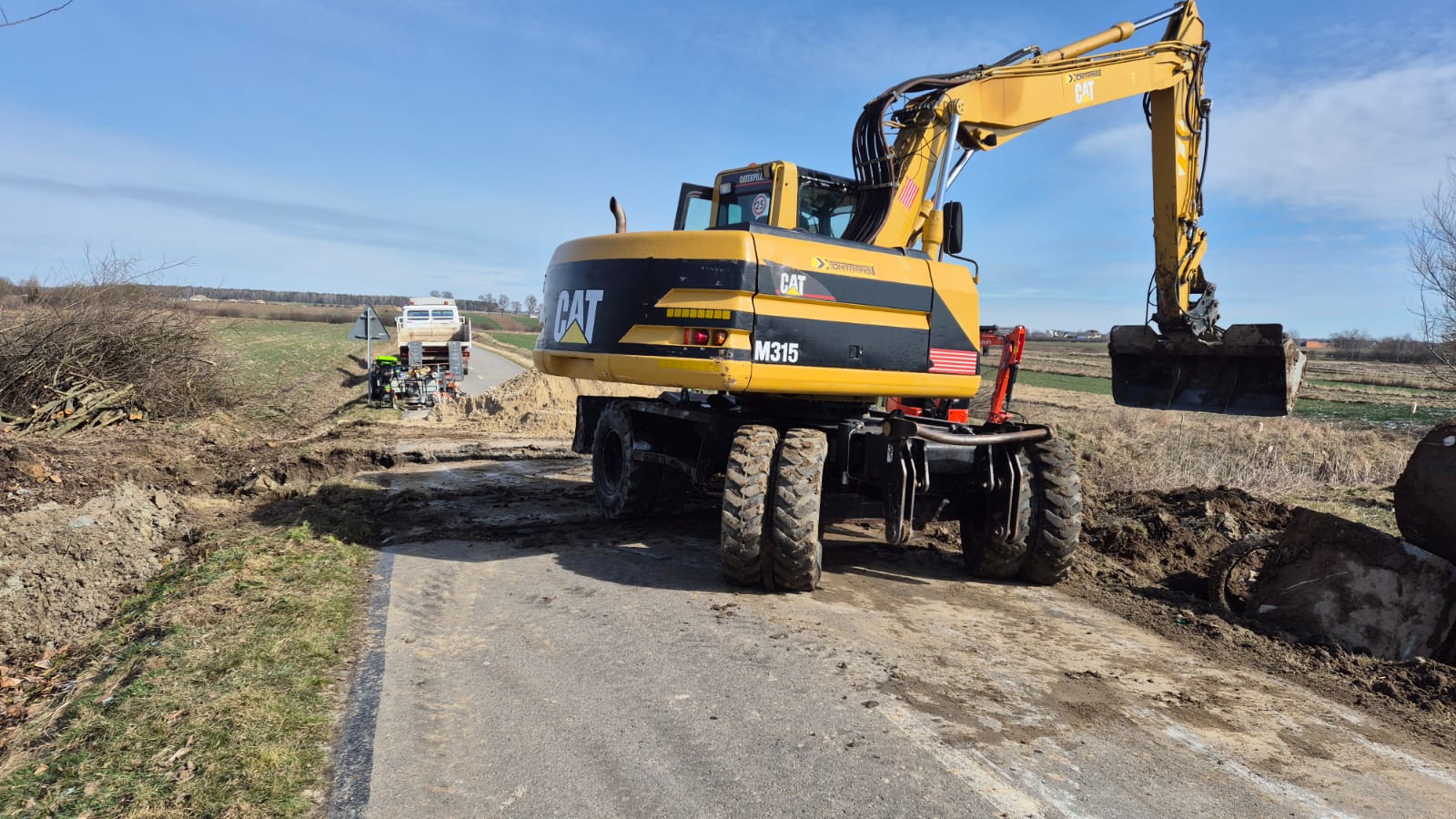 Żółto-czarny koparko-ładowarka CAT M315 na drodze z wykopanym dołem, obok stos gałęzi, w tle biały samochód i pola pod błękitnym niebem.