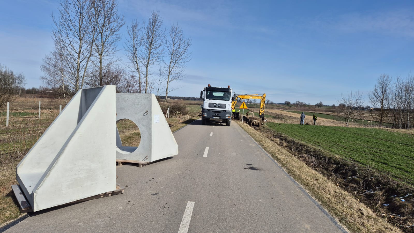 Asfaltowa droga wiejska, po lewej betonowe prefabrykaty, na drodze biały ciężarowy samochód i żółty koparko-ładowarka, po prawej dwóch ludzi na tle pól i drzew bez liści.