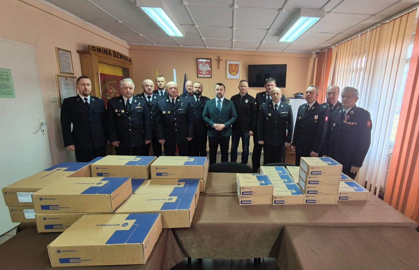 Grupa mężczyzn w mundurach i garniturach stoi za stołem z kartonowymi pudełkami Motorola w jasnym pomieszczeniu z flagami i emblematami na ścianie.