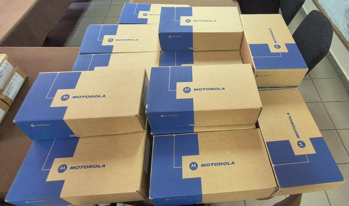 Stosy prostokątnych kartonowych pudełek w kolorze beżowo-granatowym z napisem i logo Motorola na brązowym stole, w tle czarne krzesła i jasna podłoga.