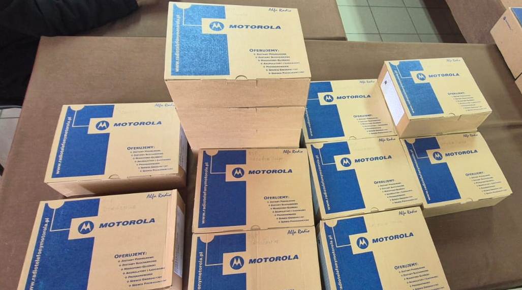 Stosy brązowych kartonowych pudełek z niebieskim logo Motorola i napisem "Oferujemy", ułożone na brązowej powierzchni.