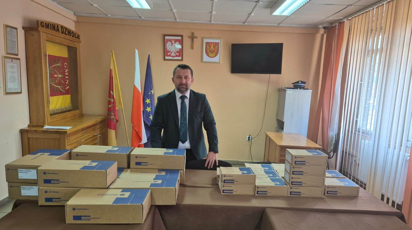 Mężczyzna w garniturze stoi za stołem z poukładanymi kartonowymi pudełkami Motorola, w tle flagi, herb Polski i herb gminy Dzwola, telewizor i krzyż na ścianie.