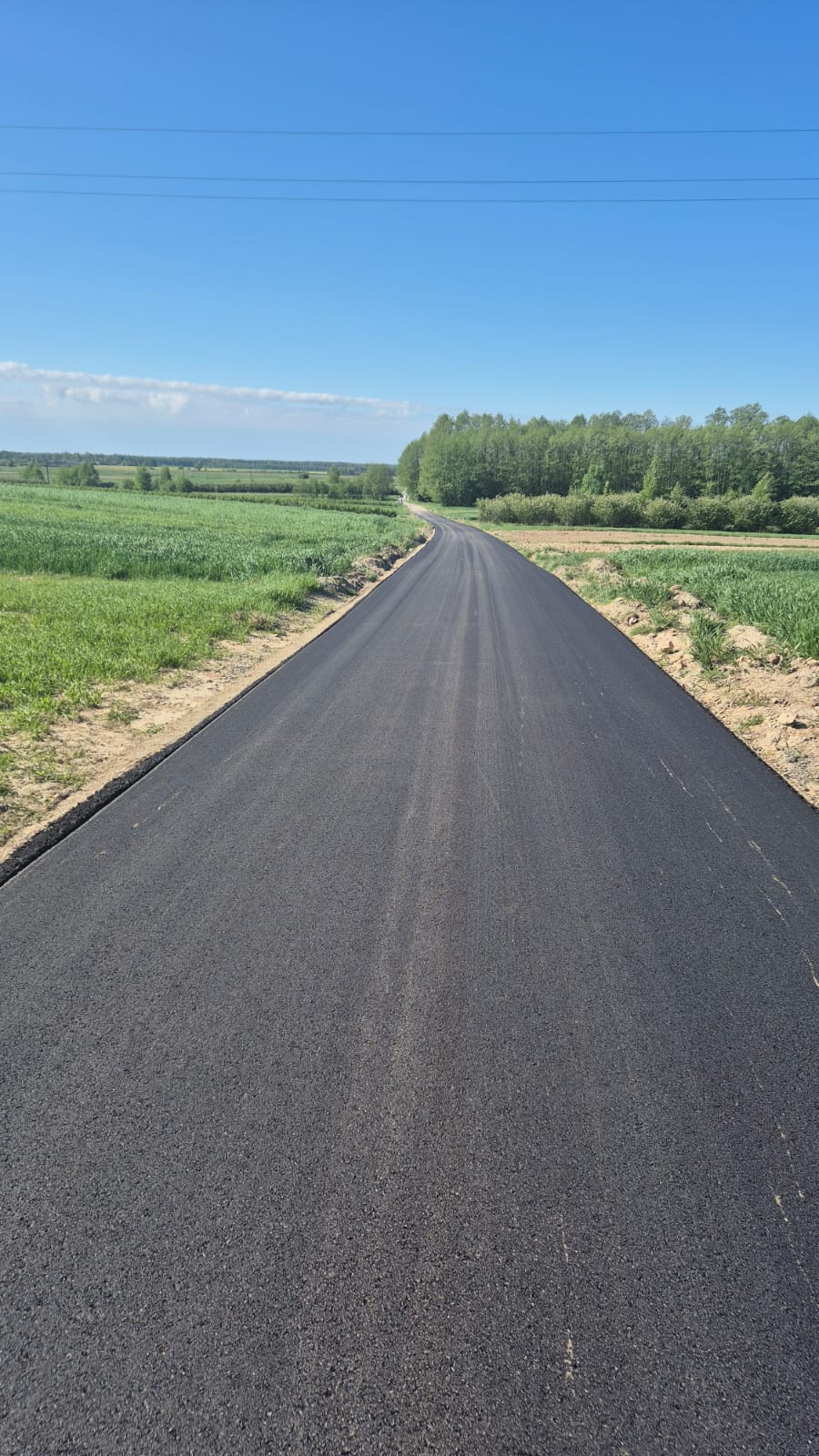 Nowa, czarna asfaltowa droga na tle zielonych pól i lasu pod jasnym, niebieskim niebem.