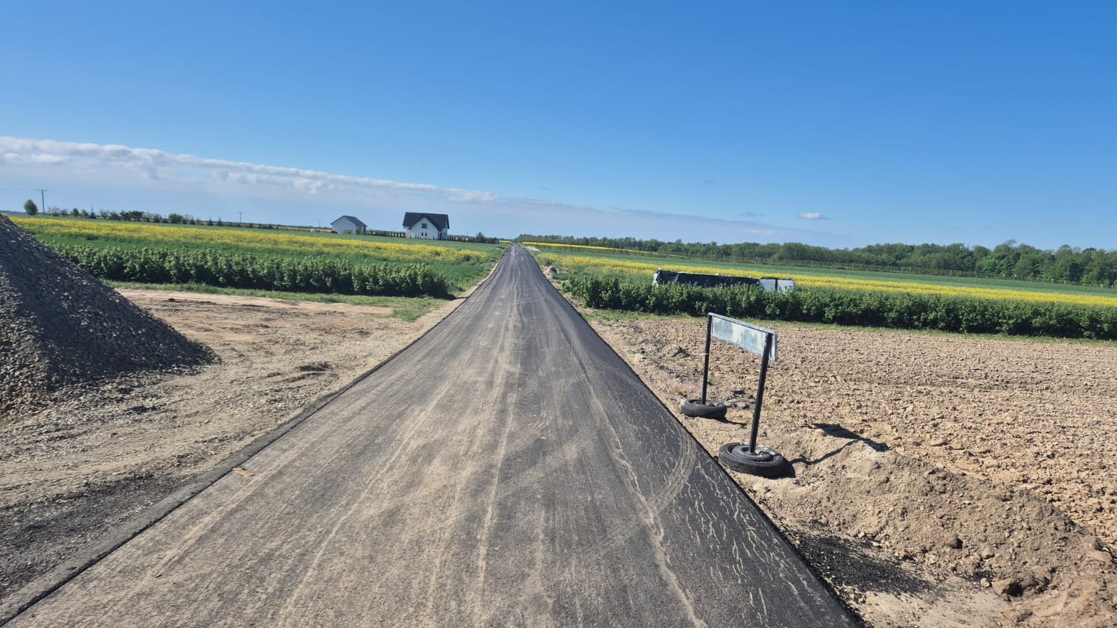 Wąska asfaltowa droga biegnąca przez pola, zielone i żółte uprawy, po lewej hałda ziemi, po prawej znak drogowy na oponach, w tle niebieskie niebo i dwa białe domy.