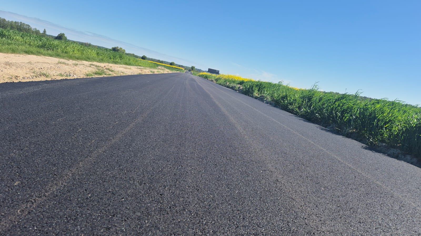 Nowa, czarna asfaltowa droga na tle zielonych traw i pól pod jasnoniebieskim, bezchmurnym niebem.