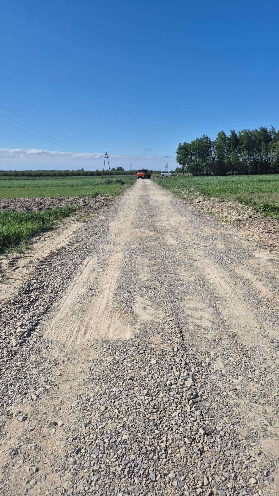 Droga utwardzona żwirem i piaskiem z zielonymi polami po bokach, na końcu droga z ciężarówką i białym samochodem, w tle drzewa i niebieskie niebo.