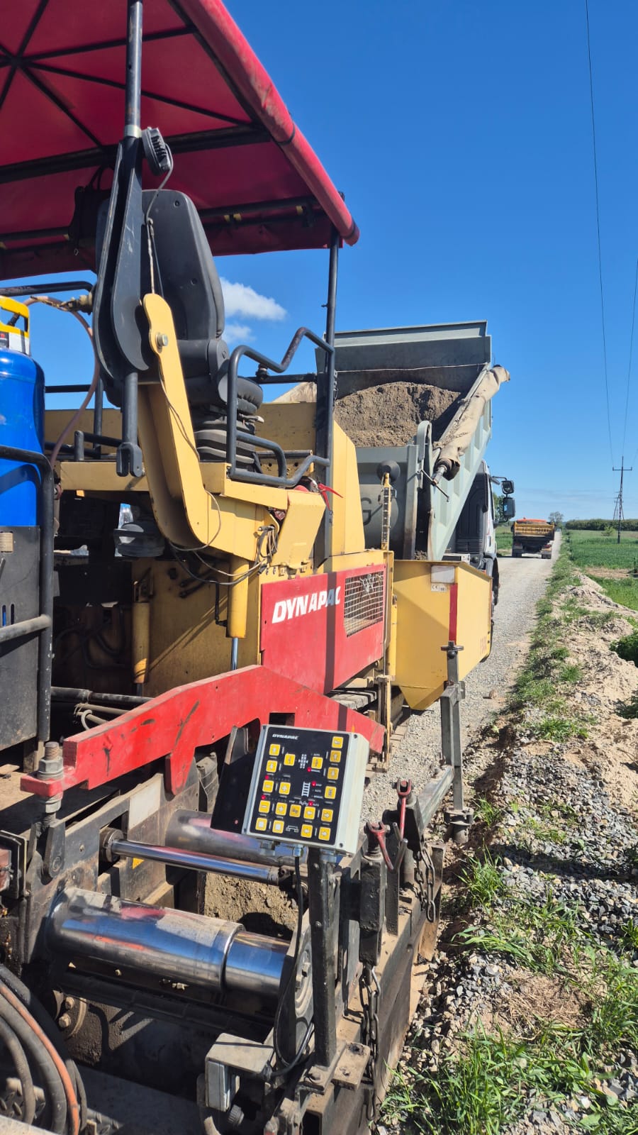 Czerwono-żółta maszyna budowlana Dynapac z panelem sterowania, siedzenie operatora pod czerwoną daszkiem, wywrotka z piaskiem na szutrowej drodze, niebieskie niebo.