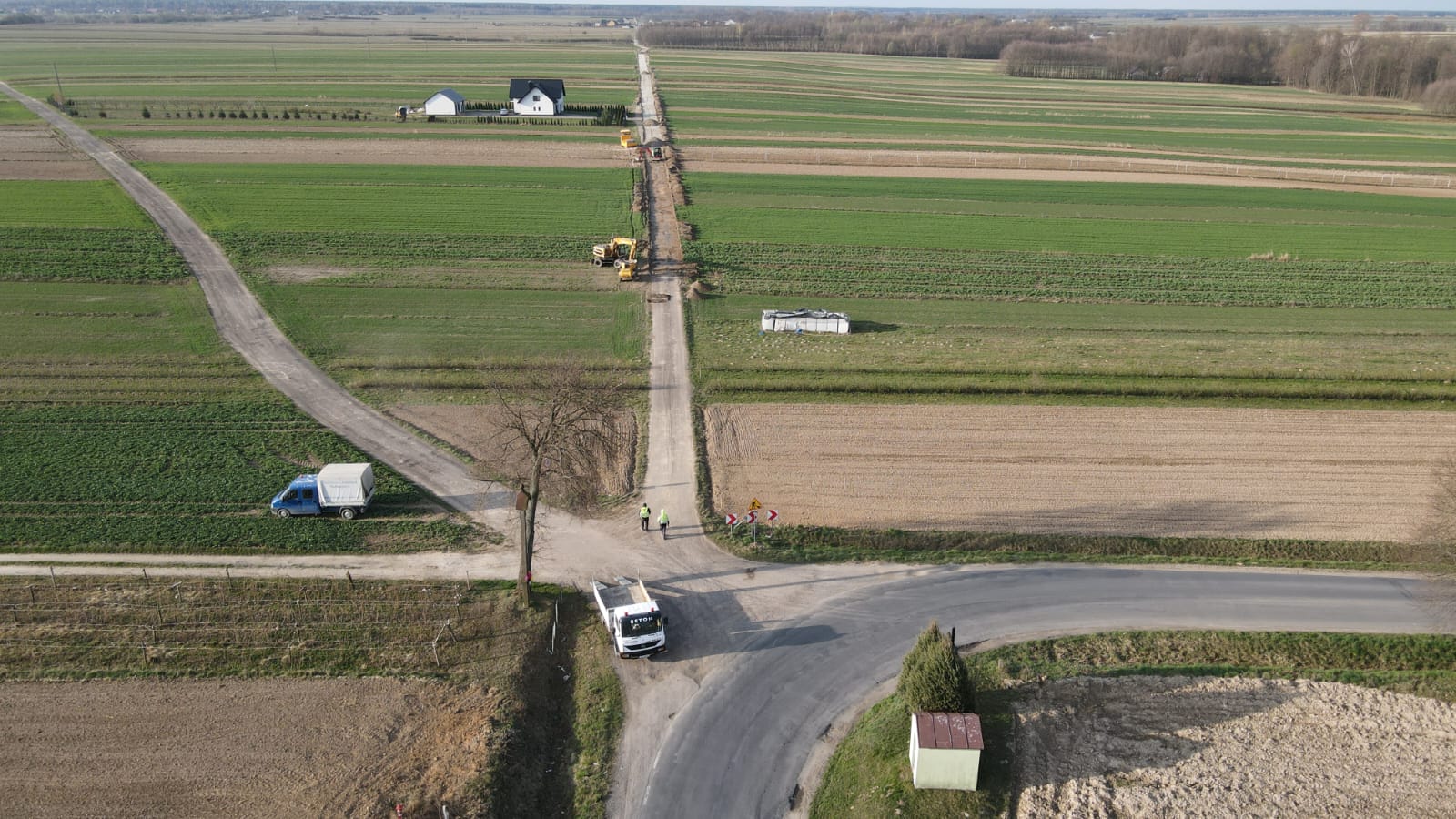 Zielone i brązowe pola uprawne, droga polna z maszynami budowlanymi, biały dom z czarnym dachem, niebieski samochód dostawczy, skrzyżowanie dróg, dwie osoby w odblaskowych kamizelkach.