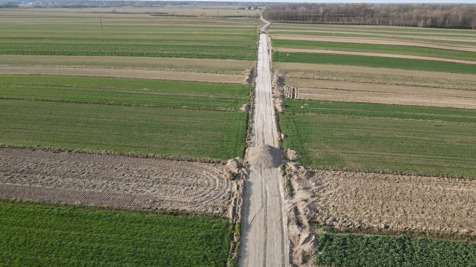 Długi, nieutwardzony, szary drogowy pas ziemi przecina pola uprawne o różnych odcieniach zieleni i brązu, widoczne ślady opon i niewielkie kopce ziemi przy drodze.