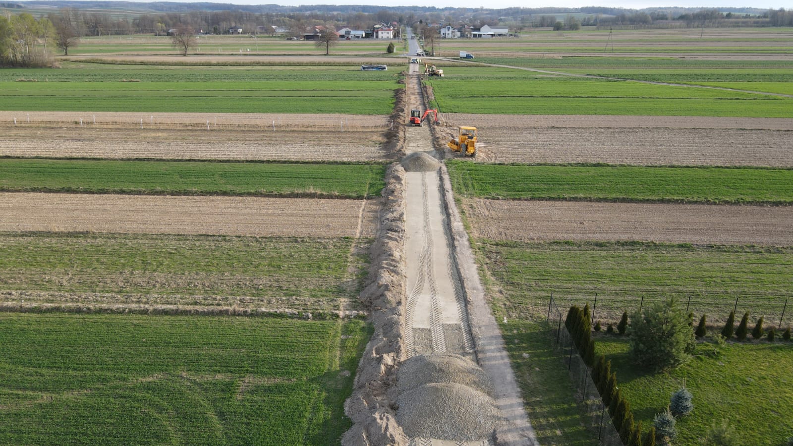 Polne drogi w trakcie budowy, żółty i pomarańczowy sprzęt budowlany, hałdy ziemi i żwiru, pola uprawne o różnych odcieniach zieleni i brązu, domy w tle na horyzoncie.