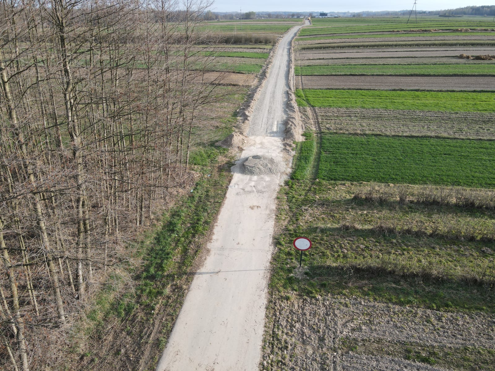 Wąska szutrowa droga z hałdą żwiru, po lewej stronie nagie drzewa, po prawej zielone i brązowe pola uprawne, przy drodze znak zakazu ruchu.