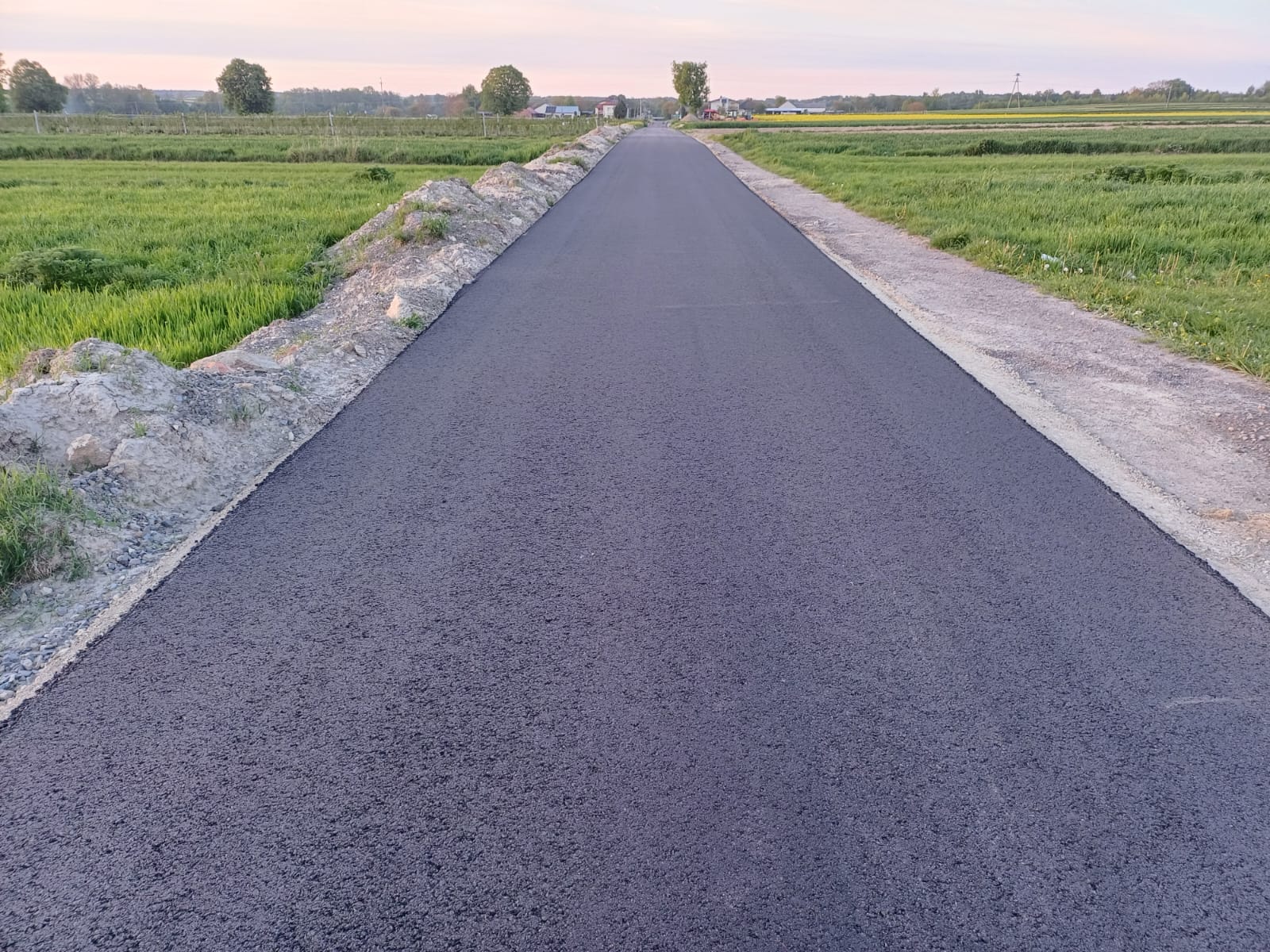 Nowa, ciemnoszara asfaltowa droga między zielonymi polami, z niewielkimi kopcami ziemi po lewej stronie, pod niebiesko-różowym niebem.