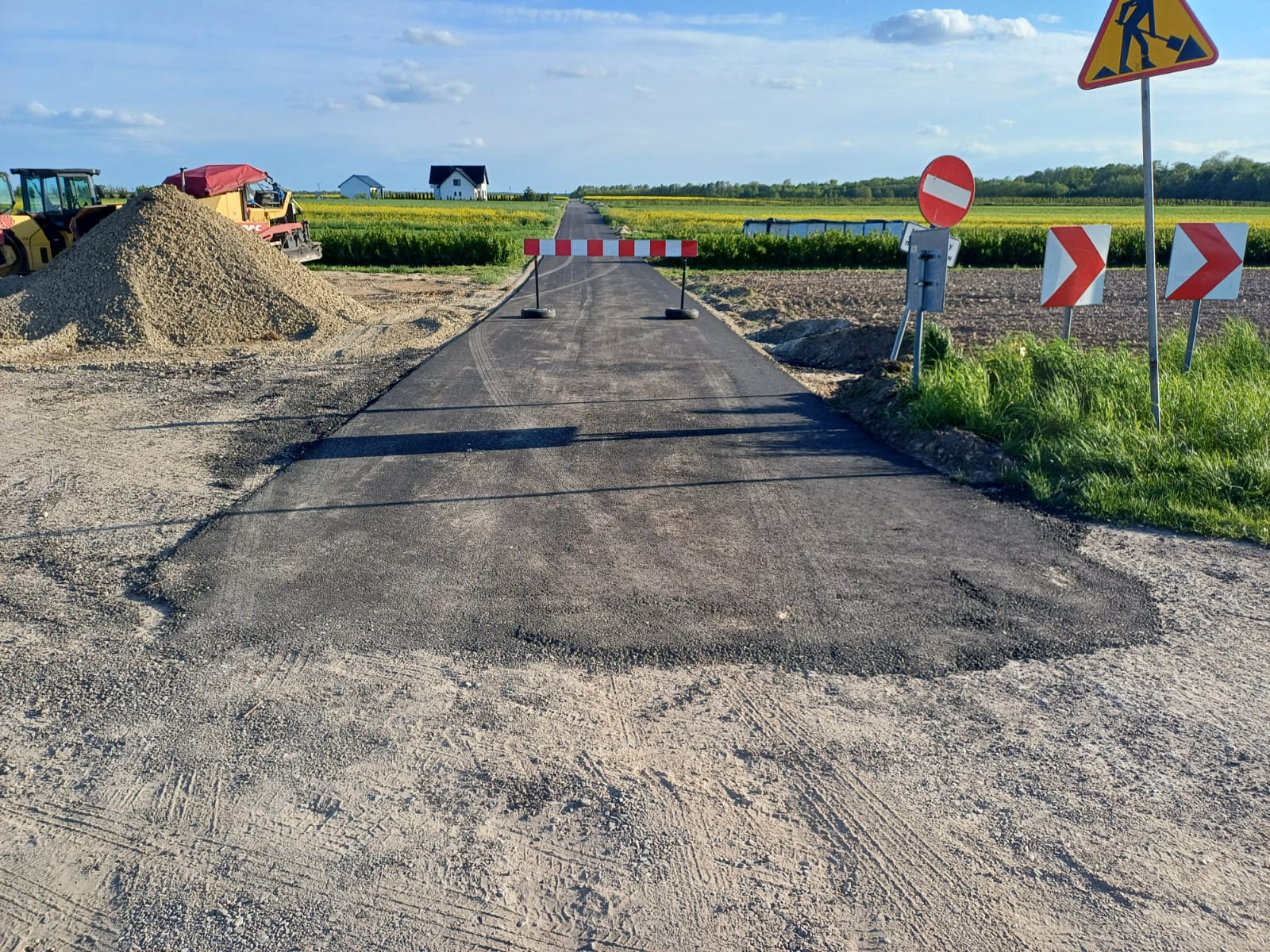 Nowa, czarna nawierzchnia drogi z barierką blokującą przejazd, obok stos żwiru i maszyny budowlane, zielone pola i znak roboty drogowe na tle niebieskiego nieba.