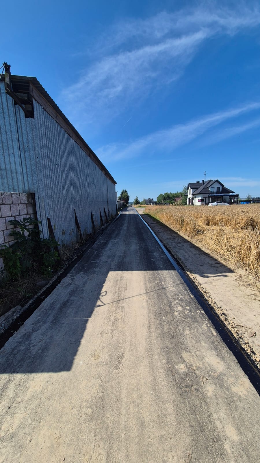Wąska, nowo wyasfaltowana droga między wysokim budynkiem z metalową ścianą a polem złotych zbóż, pod jasnym niebieskim niebem z lekkimi chmurami.