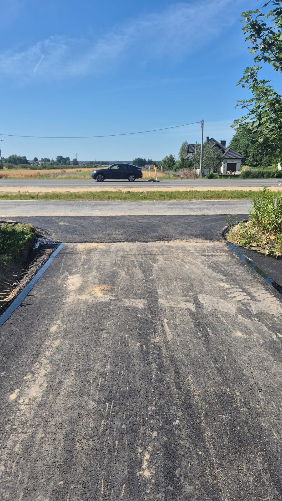 Asfaltowa droga z widokiem na czarny samochód, pola i dom z czarnym dachem pod niebieskim niebem, po bokach zielone drzewa i krzewy.