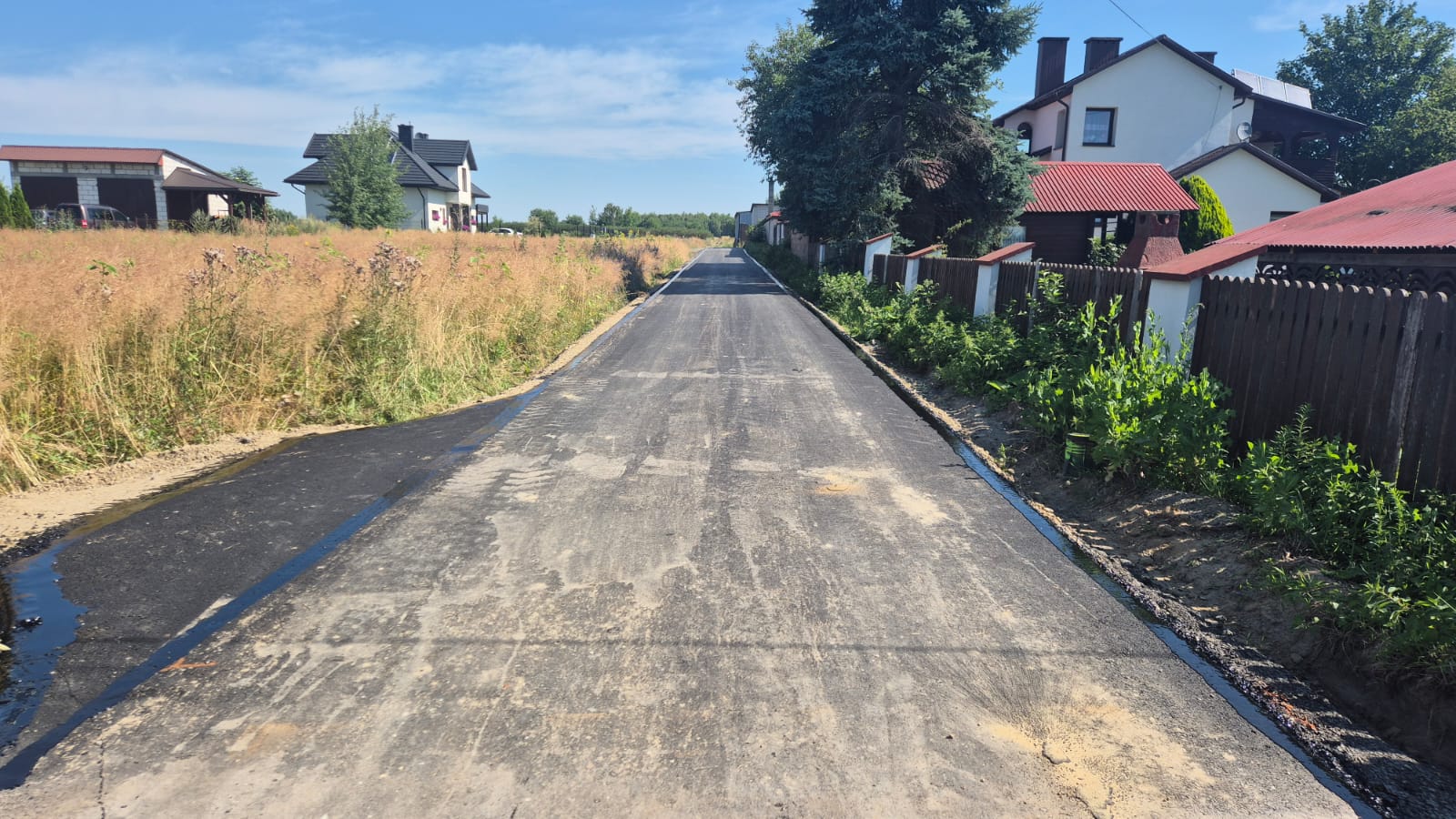 Nowa asfaltowa droga z domami po prawej stronie i suchą trawą po lewej, pod jasnym niebem.