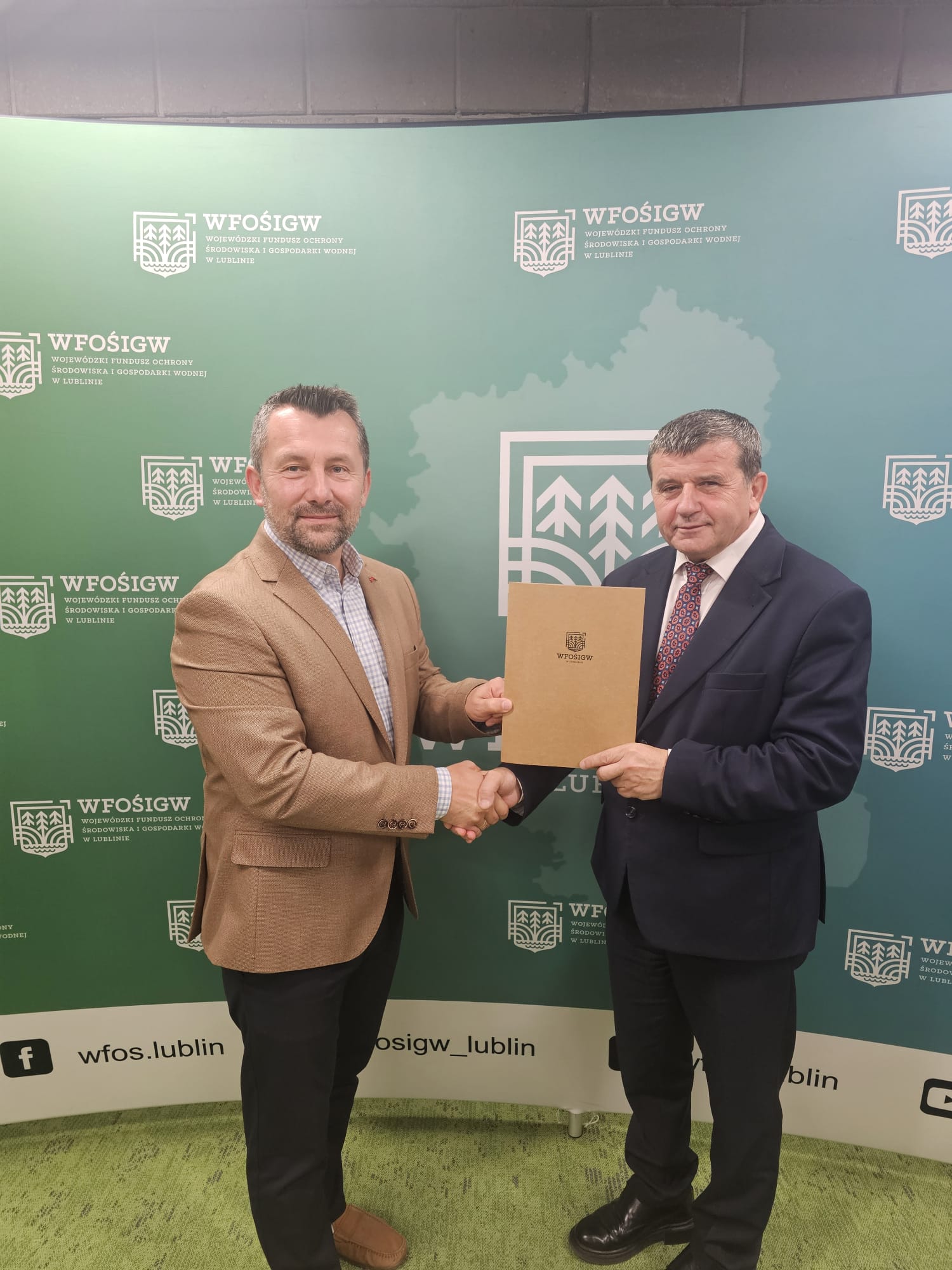 Dwaj mężczyźni uściskiem dłoni, jeden w brązowej marynarce i beżowej koszuli, drugi w ciemnym garniturze z krawatem, trzymają brązową teczkę na tle zielonego bannera z logo WFOŚiGW.
