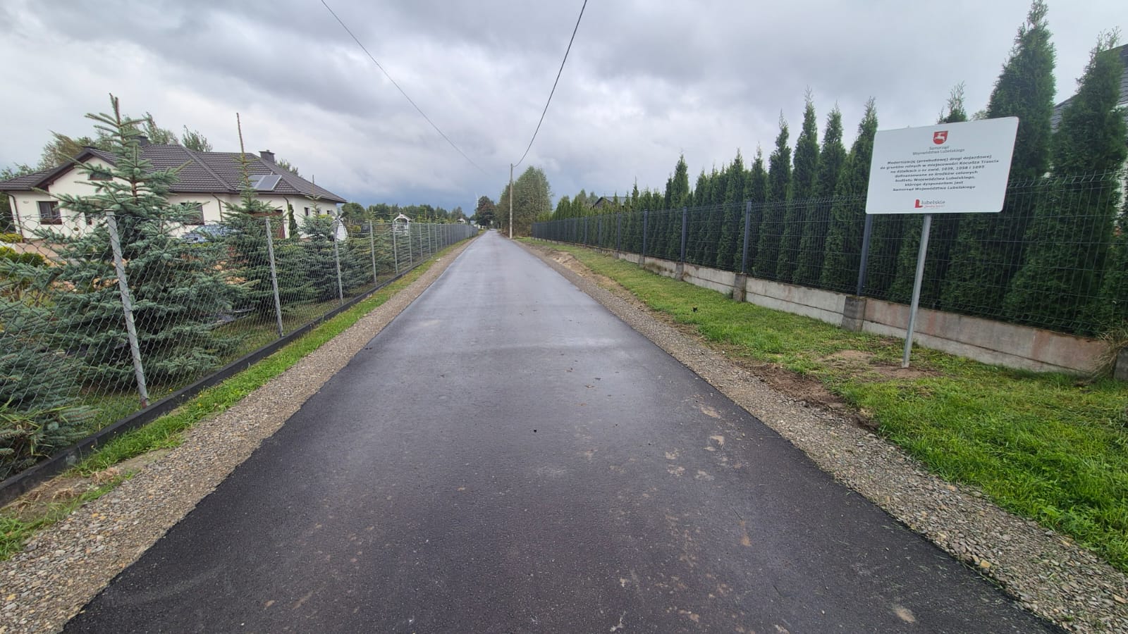 Asfaltowa droga z obu stron ogrodzona siatką, po lewej stronie dom i drzewa, po prawej gęste zielone krzewy i tablica informacyjna, pochmurne niebo.