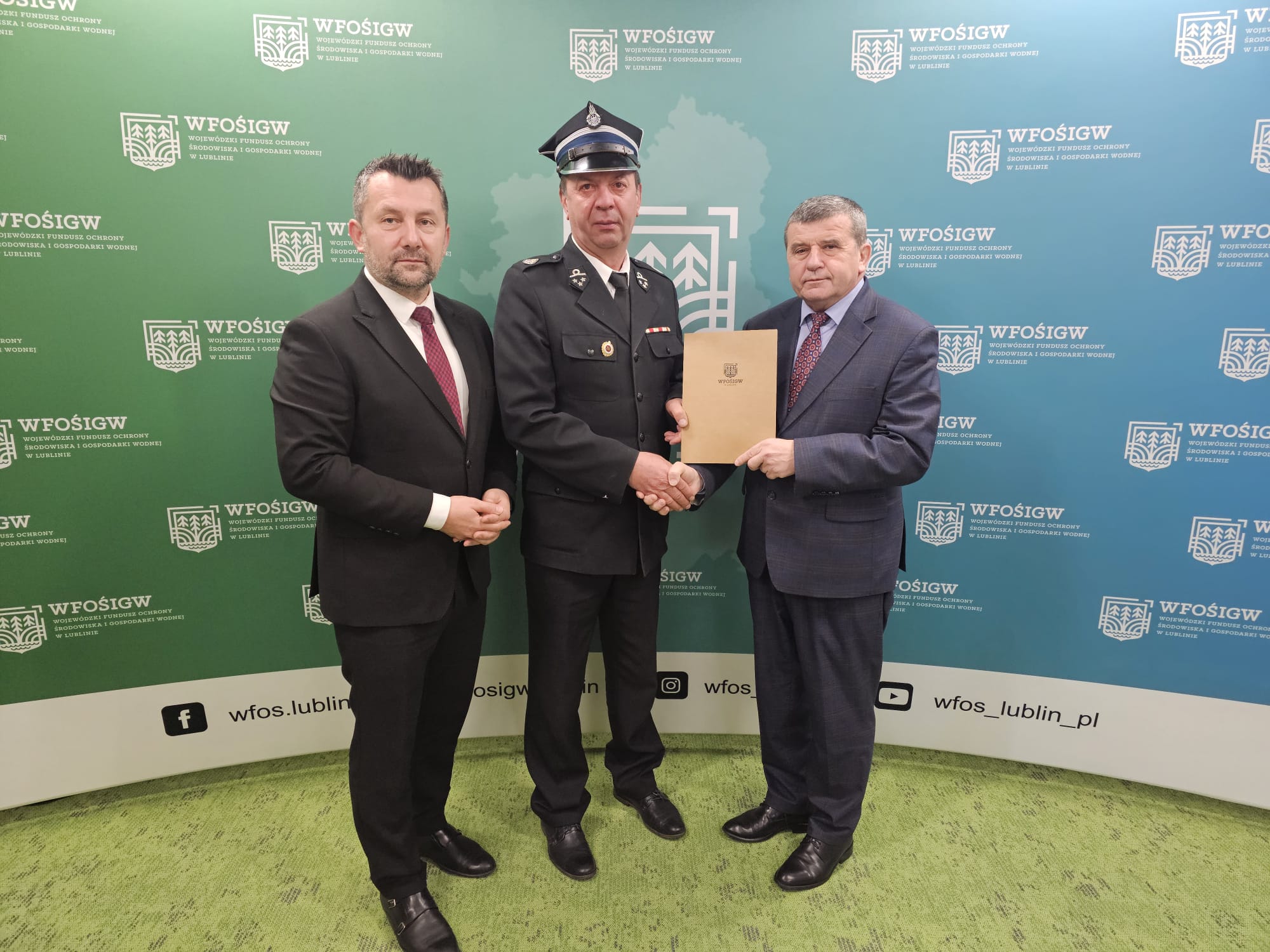 Trzech mężczyzn w garniturach, jeden w mundurze strażaka, na zielono-niebieskim tle z logo WFOŚiGW, trzymający teczkę, uścisk dłoni.