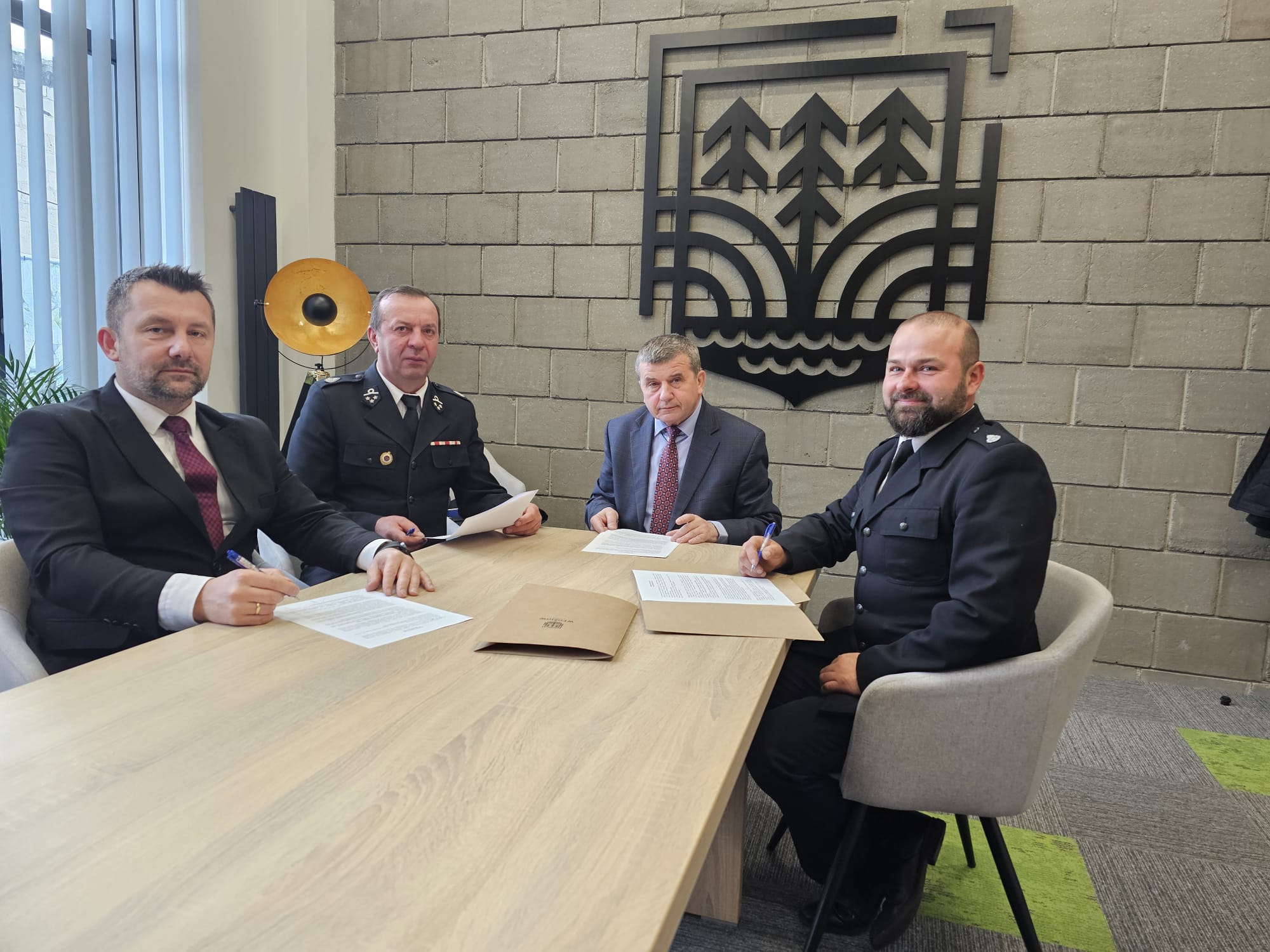 Czterech mężczyzn w garniturach i mundurach siedzi przy jasnym drewnianym stole z dokumentami, za nimi betonowa ściana z czarnym metalowym herbem i stojąca lampa z żółtym kloszem.