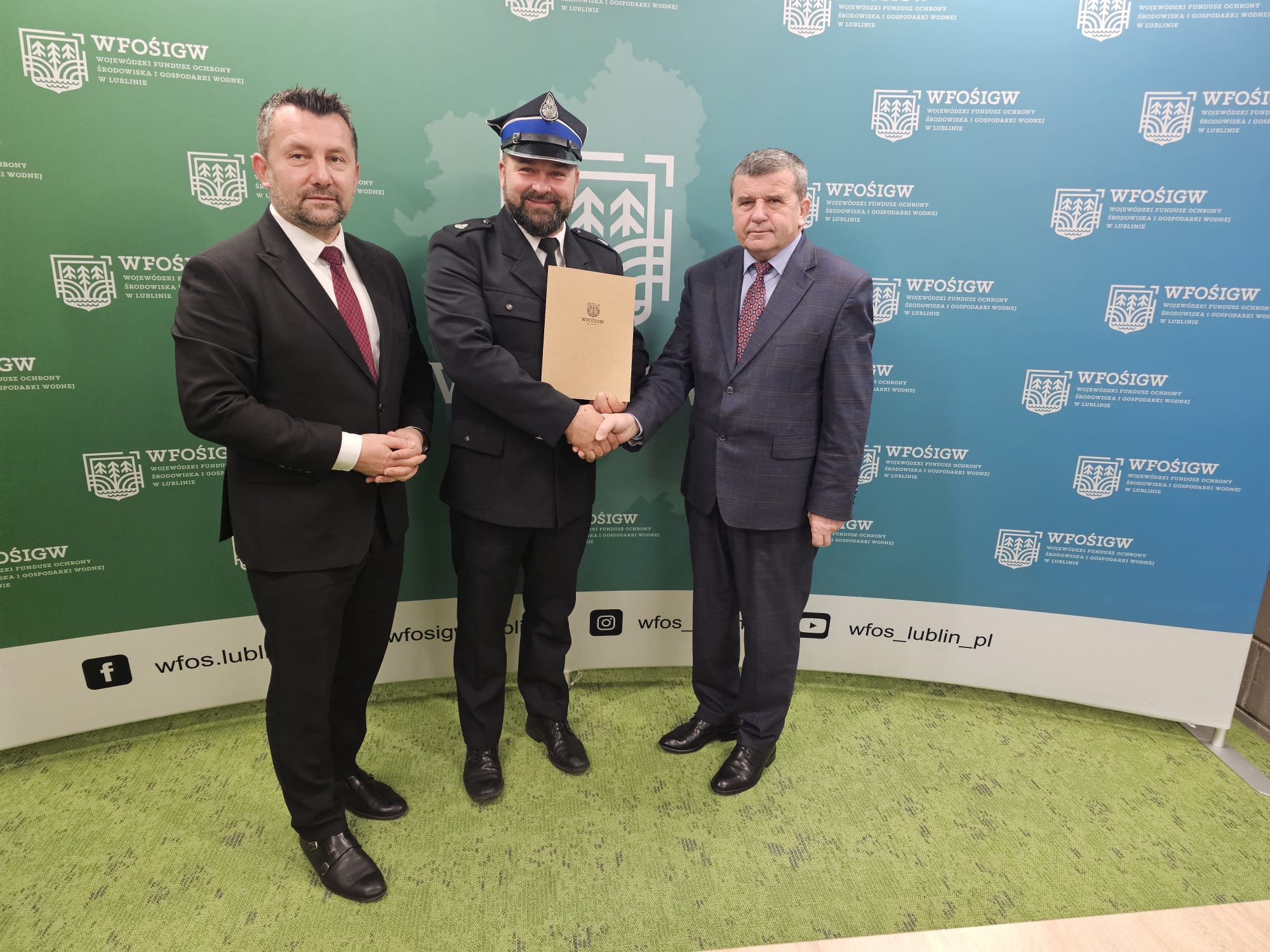 Trzech mężczyzn w garniturach na zielono-niebieskim tle z logo WFOŚiGW, środkowy w mundurze policyjnym trzyma teczkę i ściska dłoń mężczyzny po prawej.